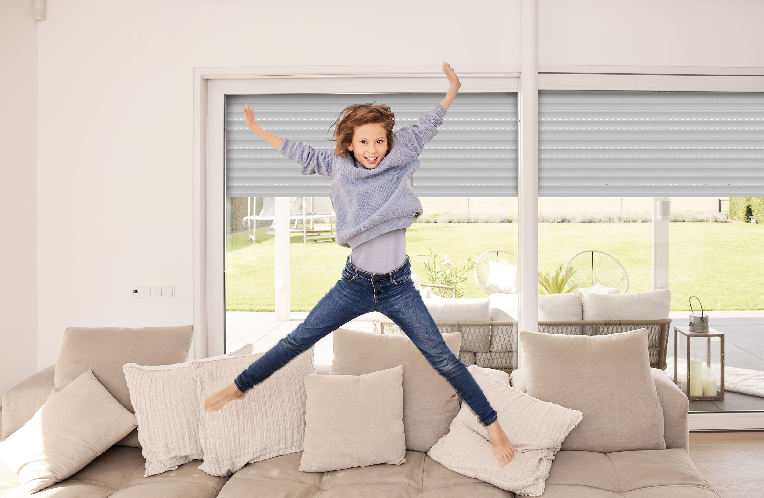 ein Mädchen springt auf der Couch im Hintergrund ist ein großes Fenster mit Rolläden